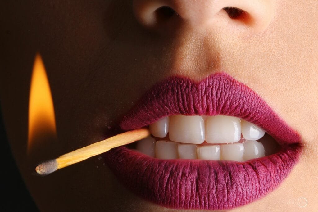 Bold close-up of lips with vivid lipstick holding a lit matchstick, creating a fierce and dynamic image.